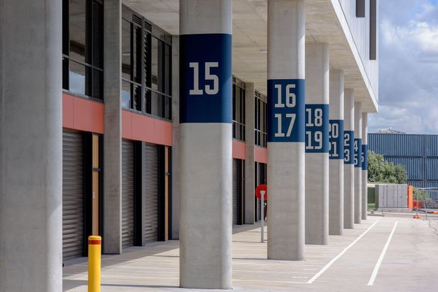 Exterior view of a contemporary commercial or industrial building with a long covered colonnade. Large cylindrical concrete columns are marked with oversized white numbers on dark blue bands, while red horizontal facade panels, tall glazed openings, roller doors, and painted vehicle parking lines define the frontage. The scene has a clean, functional, modern architectural character.