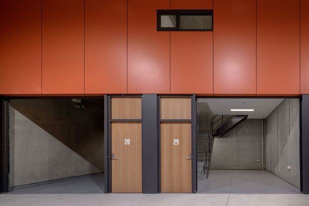 A contemporary commercial or institutional interior with a bold burnt-orange wall panelled across the upper level. Two timber-look doors numbered 28 and 29 sit within dark metal framing, flanked by exposed concrete walls and a stairwell leading upward. The space has an industrial, utilitarian character with a polished concrete floor, grey concrete surfaces, black metal details, and a narrow horizontal window.