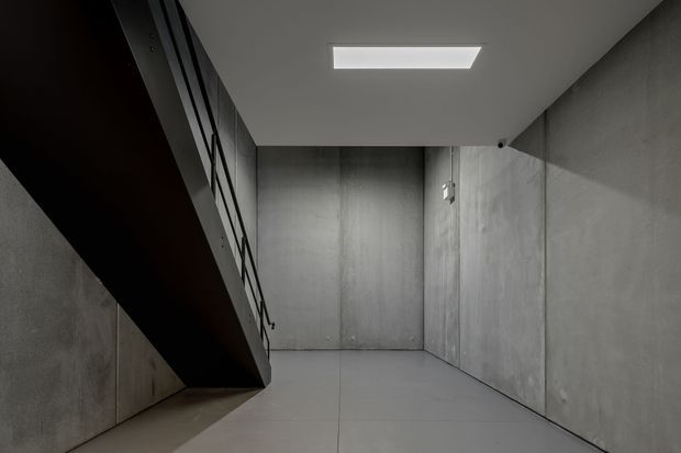 A stark minimalist concrete interior corridor or landing beneath a dark angular stair or mezzanine structure. Smooth grey concrete walls and floor create a monolithic, industrial atmosphere, with a simple rectangular ceiling light and black metal railing visible along the upper structure.