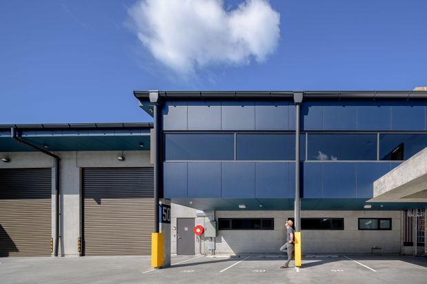 Exterior view of a contemporary commercial or industrial building with blue-grey metal-clad upper walls, broad dark-framed glazing, roller garage doors, loading or service-bay access, concrete surfaces, and a projecting flat-roof canopy. The facade is photographed under a clear blue sky.