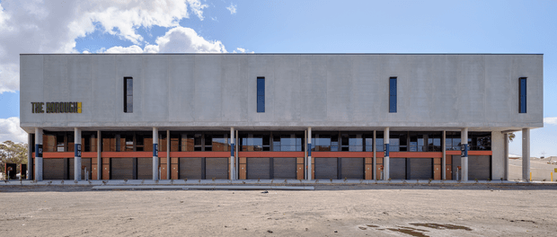 Front-on exterior photograph of a long, rectilinear multi-storey commercial building. The upper level is a plain grey concrete or rendered facade with narrow vertical windows, while the lower level has repetitive open bays, columns, dark glazed or screened sections, and warm reddish-brown panels. Large paved forecourt in front, with blue sky and clouds in the background. A building sign is visible at the left side of the facade.