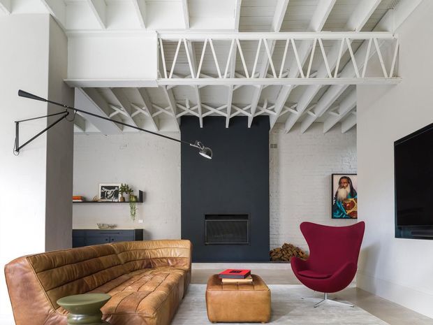 Bright contemporary living room with exposed white-painted roof trusses and beams, white brick walls, a dark charcoal fireplace feature wall, and pale flooring. The room contains a cognac leather sofa and ottoman, a sculptural red lounge chair, a wall-mounted television, track-style articulated lighting, and restrained decorative accents.
