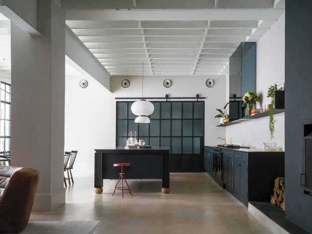 Open-plan industrial-style kitchen and living area with a dark island and cabinetry, black-framed glazed partition doors, white exposed ceiling beams, concrete flooring, pendant lighting, and a built-in fireplace surround. A partial living room and dining area are visible at the left.