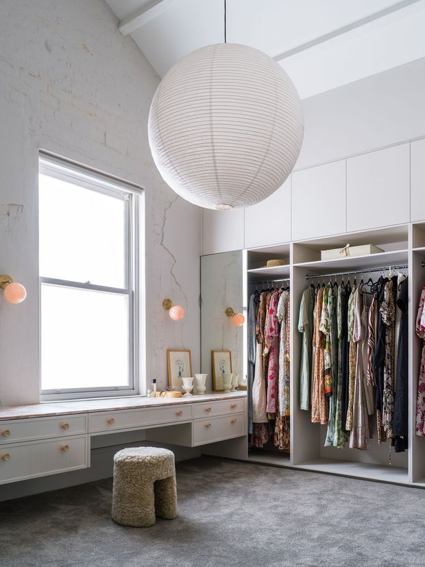 Bright, refined dressing room with extensive built-in white cabinetry, open hanging rails filled with clothing, drawers beneath a window, and a small textured stool on grey carpet. A large ribbed paper pendant light hangs from a white ceiling, while exposed pale brick and softly coloured wall sconces add character.