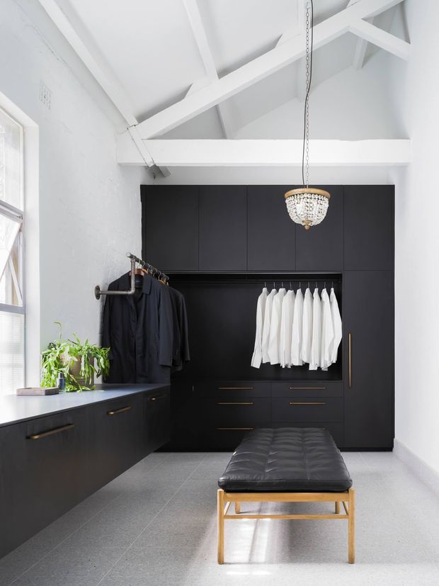 Bright, compact walk-in wardrobe with dramatic matte-black built-in cabinetry, open hanging rails displaying dark and white garments, integrated drawers, and a black upholstered bench with timber legs. White-painted walls and exposed pitched ceiling beams contrast with the dark joinery, while a decorative pendant light and a small plant add character. Natural light enters through the side window.