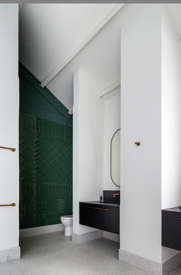 A contemporary bathroom with a dramatic dark green glazed tile feature wall in a herringbone and vertical pattern, white walls and sloped ceiling, grey terrazzo-style flooring and skirting, and matte black floating vanities with brass hardware. A white toilet and rounded-edge mirror are visible, along with a partial adjacent vanity.