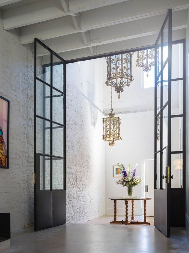 Bright loft-like interior with exposed white-painted timber ceiling beams, white-painted brick walls, polished concrete flooring, and tall black steel-framed glazed doors. Ornate antique-style pendant chandeliers hang above a small timber console table with flowers, creating a dramatic contrast between industrial architectural elements and decorative traditional detailing.