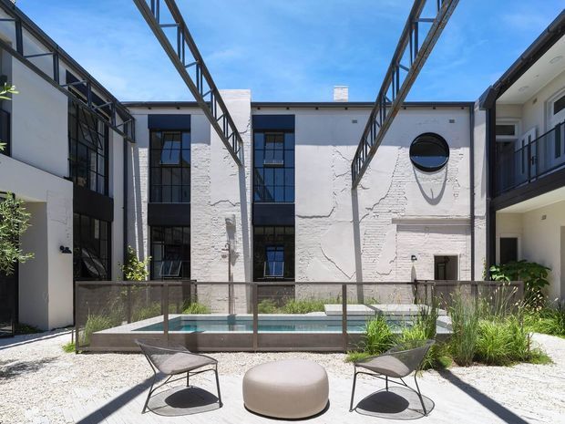 A bright contemporary courtyard surrounded by multi-storey buildings with white textured masonry walls, large black-framed windows, balconies, and exposed overhead steel framing. The landscaped central space includes a narrow reflective pool, gravel paving, ornamental grasses, and two outdoor lounge chairs with a rounded ottoman. Strong sunlight creates crisp shadows, giving the courtyard an airy, modern architectural character.