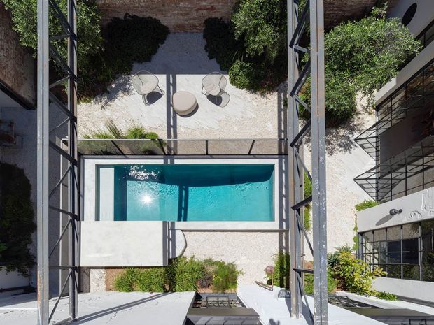 Aerial view into a modern courtyard enclosed by a multi-level building, featuring a rectangular turquoise swimming pool, pale gravel landscaping, sculpted shrubs, outdoor lounge seating, and dark steel-framed glazing and walkways. The composition highlights the relationship between the pool, courtyard garden, and surrounding architecture.