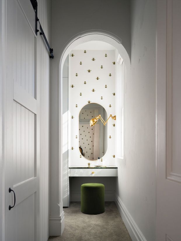 A narrow, bright dressing or vanity area viewed through white paneled doors. The space features an arched opening with decorative trim, a floating white vanity with a dark stone or glass top, an oval mirror, brass wall-mounted lighting, patterned wallpaper with small gold motifs, white wall panelling, carpeted flooring, and a small green upholstered stool.