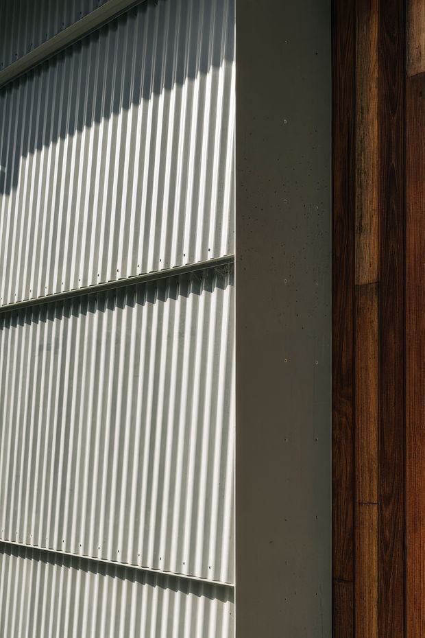 Close-up architectural detail showing vertically ribbed corrugated metal cladding, a dark smooth structural panel or column, and adjoining dark timber wall cladding. Strong directional sunlight creates pronounced shadows across the textured metal surface. No room context or furnishings are visible.