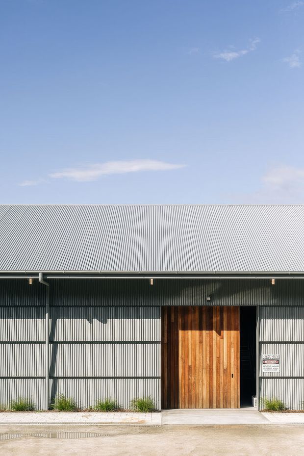 Front-facing exterior view of a simple industrial-style building with a broad corrugated metal gable roof and matching vertically ribbed metal facade. A large warm timber-clad entrance door is set into the facade beside a dark open doorway. Concrete paving and sparse planted grasses line the building, with a clear blue sky above.