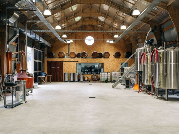 A spacious industrial distillery or brewery interior with polished stainless-steel processing tanks, copper vessels, hoses, barrels, and production equipment. The building features a high timber-framed pitched roof with exposed rafters, exposed galvanized ventilation ducts and services, pendant lighting, concrete flooring, and warm plywood wall cladding. A central service counter and barrel display sit at the rear.