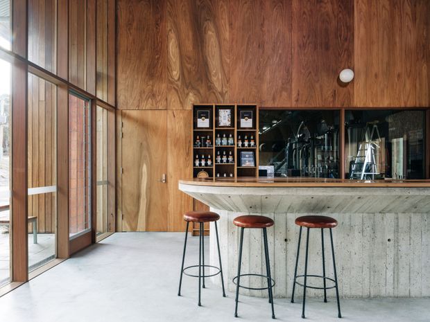 A refined bar area in a contemporary hospitality-style interior, featuring a full-height dark timber wall and matching glazed timber-framed doors on the left. A concrete-fronted bar with a pale stone or concrete countertop spans the right side, with open shelving displaying bottles and glassware behind it. Three slim black metal bar stools with brown leather seats line the counter. The space is well lit by tall glazing and has a polished, industrial-leaning material palette.