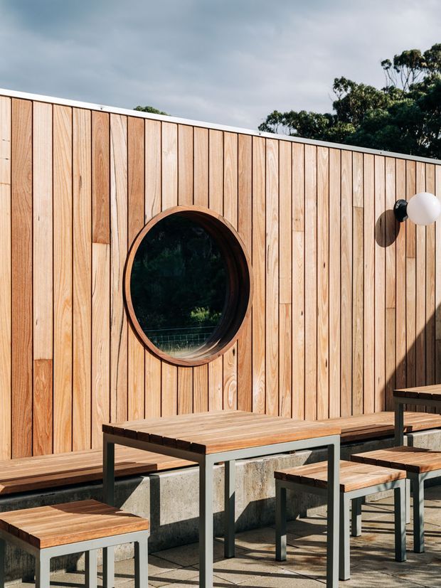 Sunny outdoor terrace or courtyard with a vertical timber-clad privacy wall, a round porthole-style window, integrated timber bench seating, and a simple timber-and-metal table with stools. The space has a warm, contemporary character and overlooks leafy vegetation beyond the wall.