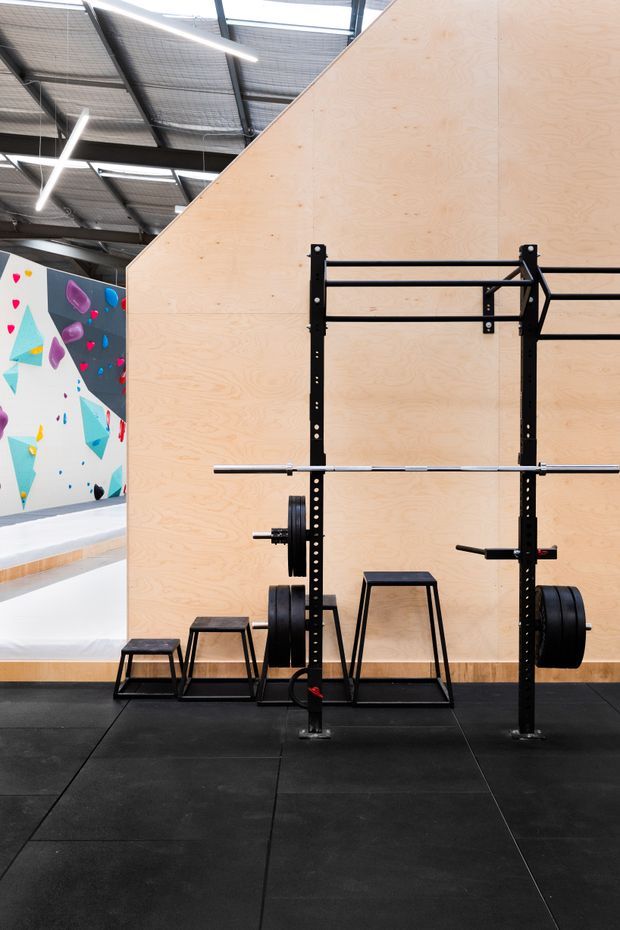 A commercial-style gym interior with a black power rack, barbells, weight plates and workout benches positioned against a large pale plywood partition wall. The space has black rubber flooring, an exposed industrial ceiling with linear strip lighting, and colourful climbing-wall graphics visible in the background.
