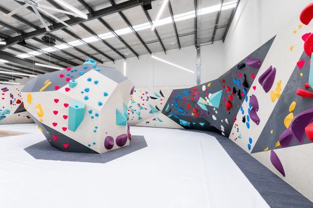 A bright, expansive indoor climbing gym with large geometric climbing walls and bouldering volumes covered in colourful climbing holds. The space has a high exposed metal roof structure, white walls and floor, linear strip lighting, and bold red, blue, purple, yellow, and teal accents.