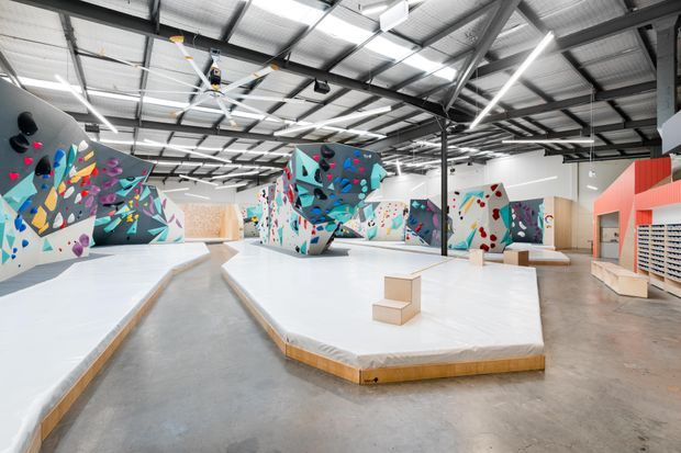 Large indoor climbing and bouldering gym with a high industrial warehouse ceiling, exposed steel structure, corrugated metal roofing, bright linear lighting, polished concrete floors, and numerous vividly painted climbing walls and padded platforms. The space is open, expansive, and brightly lit, with strong geometric forms and colourful climbing holds.