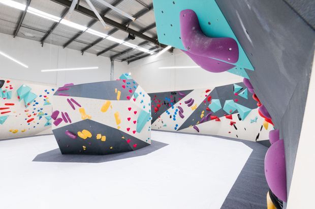 An indoor climbing gym with angular climbing walls and bouldering structures covered in colorful handholds and footholds. The space has a bright white floor, exposed industrial steel roof framing, linear strip lighting, and vivid geometric graphics in turquoise, magenta, yellow, red, blue, and grey.