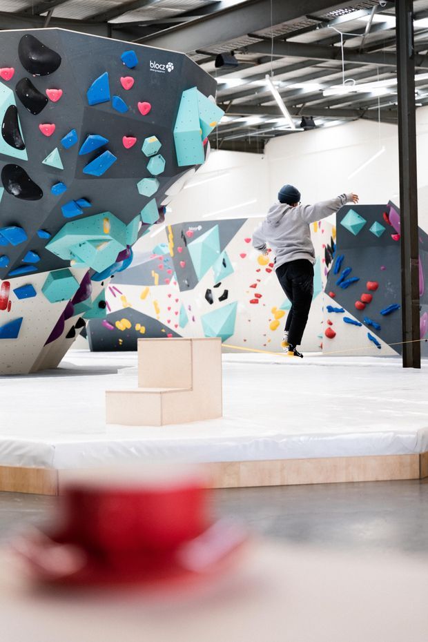 Interior view of a commercial indoor bouldering and climbing gym. The space features large angular climbing walls covered with colourful climbing holds, padded white flooring, a timber-edged raised landing, exposed steel roof structure, and bright linear industrial lighting. A climber is visible in the background.