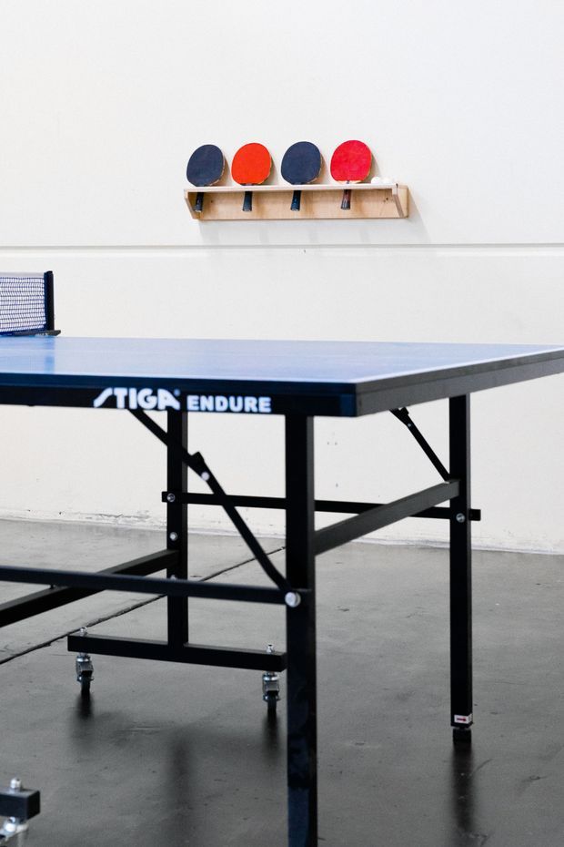 A close-up product-style photograph of a foldable table tennis table in a clean, sparse indoor space. The blue playing surface and black metal support frame dominate the foreground, with a wall-mounted wooden rack holding red and dark-blue table tennis bats and accessories in the background. The room has white walls and a smooth grey concrete floor.