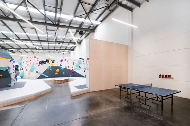 Bright industrial-style indoor recreation facility combining a table-tennis area with a colourful climbing and bouldering gym. The large open space has polished concrete flooring, exposed steel roof framing and services, white walls, plywood-clad partitions, linear LED lighting, and vibrant climbing walls with padded platforms.