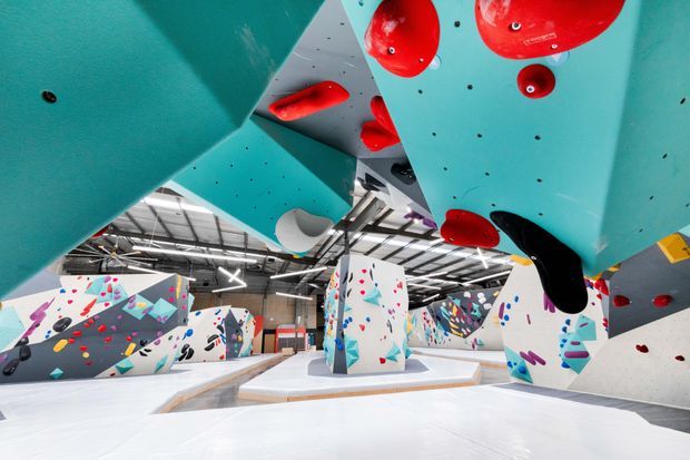 A bright indoor bouldering and climbing gym with extensive angular climbing walls, overhanging turquoise and white structures, colourful climbing holds, padded white flooring, and an exposed industrial ceiling. The image emphasizes the climbing infrastructure and open recreational space.