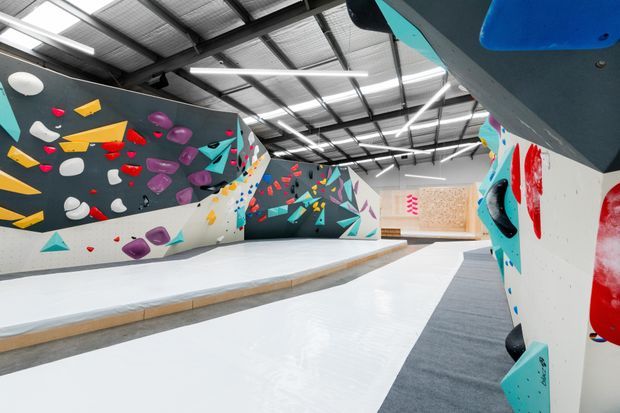 Wide interior view of an indoor climbing gym with multiple sculpted climbing walls, colourful handholds and footholds, geometric painted graphics, padded flooring, and an exposed metal industrial ceiling with linear lighting.
