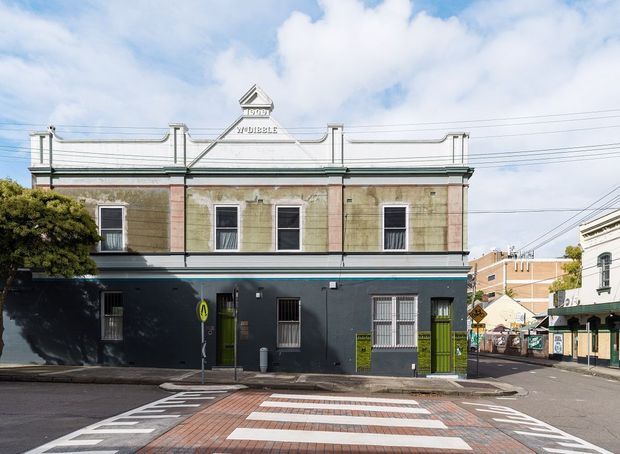 Street-facing view of a two-storey heritage commercial building on a corner site. The facade combines a dark painted ground floor with an ornate rendered upper level, tall sash-style windows, decorative parapet detailing, and a small central gable bearing lettering. A zebra crossing and surrounding urban streetscape are visible in the foreground.