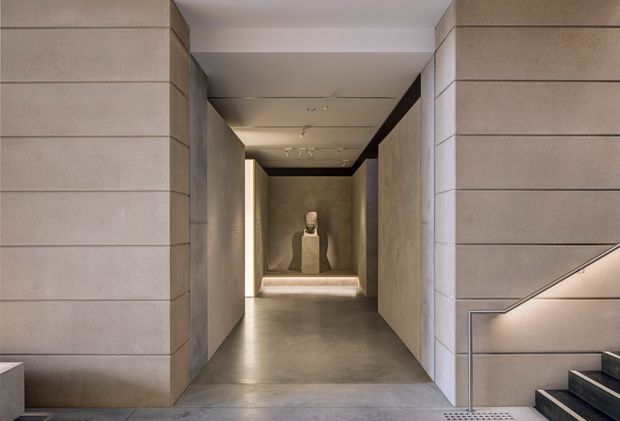 A refined contemporary interior passage or gallery with warm grey stone or concrete wall panels, polished concrete flooring, recessed linear lighting, and a sculptural display at the far end. A stairway with dark treads and an understated metal handrail is visible on the right. The composition is minimalist, symmetrical, and softly illuminated.