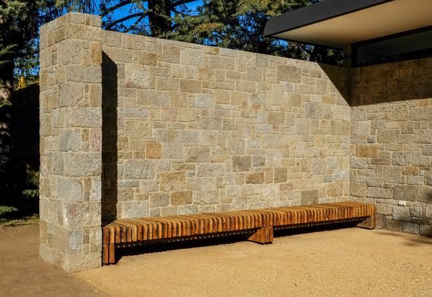 Exterior architectural detail showing a long built-in timber bench set against a textured natural stone masonry wall. The courtyard-like area has a sandy ground surface, a projecting flat roof or canopy at upper right, and dark-framed glazing beneath it. Mature trees are visible beyond the wall.