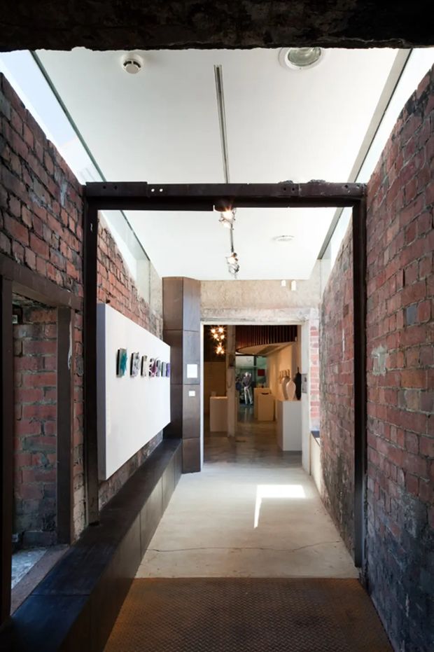 A narrow industrial-style interior passage with exposed red brick walls, dark steel framing and beams, a polished concrete floor, and a white ceiling with linear track lighting. A large white display panel with artwork is mounted along the left wall, leading toward a brighter gallery-like space with display plinths and sculptural objects.