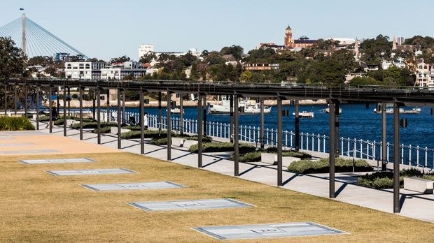 A landscaped waterfront promenade with a broad lawn, rectangular inset metal panels, a covered pedestrian structure with dark steel posts, and a white metal balustrade along the blue harbour. A cable-stayed bridge and dense urban buildings are visible across the water.