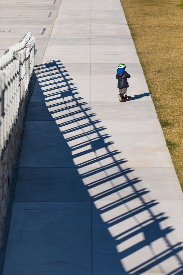 A long pale concrete pedestrian path runs beside a white metal fence, whose repeated vertical rails cast strong geometric shadows across the pavement. A small child wearing a helmet rides a scooter near the upper right, with a strip of dry grass alongside the path. The image has a high-angle perspective and strong light-and-shadow contrast.