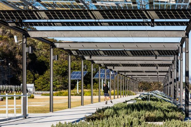 A long covered outdoor walkway lined with steel columns and an extensive solar-panel canopy. The path runs beside landscaped planting and a dry grassy open area, with additional solar panels and trees in the background. The image presents a contemporary sustainable site structure with strong repetitive geometry and integrated photovoltaic roofing.