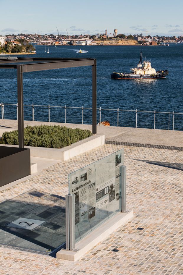 A waterfront public plaza or promenade overlooking a busy harbour, with a tugboat on the water and an urban shoreline beyond. The foreground features patterned stone paving, low landscaped planters, glass interpretive signage, metal railings, and a dark steel canopy or pergola structure. The scene is bright, open, and contemporary, with strong views across the water.
