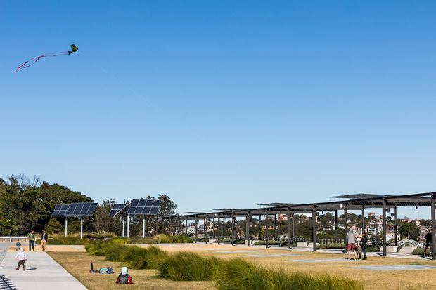 An expansive landscaped outdoor public space under a clear blue sky, featuring solar-panel canopies, open steel-framed shelter structures, paved paths, ornamental grasses, and distant urban buildings. People and recreational activity are visible, including a kite in the sky.