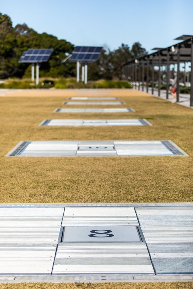 Outdoor landscaped area with a row of rectangular metal-covered ground hatches or service access panels set into dry grass, including a foreground panel marked with the number 8. Solar-panel canopies stand to the left, while a long open-sided covered structure and walkway extend along the right. The image is photographed at ground level with shallow depth of field and strong linear perspective.