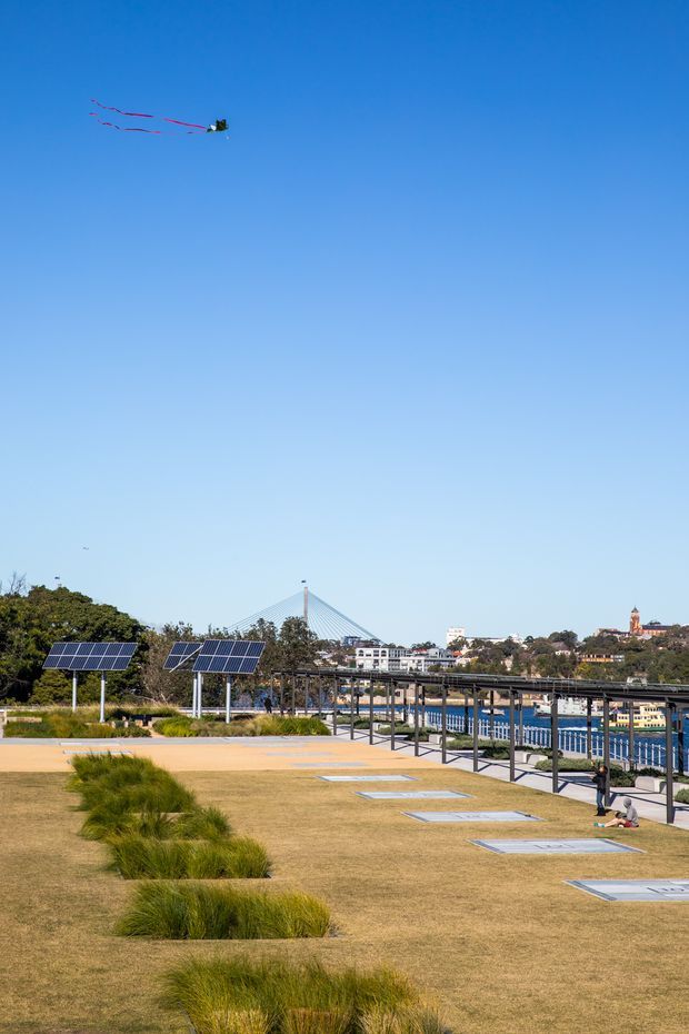 An open waterfront public landscape with a broad manicured lawn, native ornamental grass planting, rows of ground-level rectangular panels or skylight-like features, solar panel arrays, and a fenced pedestrian promenade beside the water. A suspension bridge and urban buildings are visible in the background beneath a clear blue sky, with a kite overhead.
