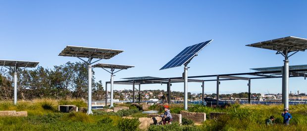 A landscaped outdoor site featuring an array of elevated photovoltaic solar panels mounted on steel support columns and frames. Native grasses, trees, large rocks, and people occupy the foreground, while low-rise buildings and a waterfront or urban landscape are visible in the distance under a clear blue sky.