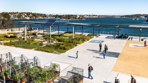 A landscaped waterfront public recreational space beside a broad bay, featuring paved courts and open gathering areas, solar-panel shade structures, native planting beds, and community garden plots in the foreground. Residential development and boats are visible across the water.