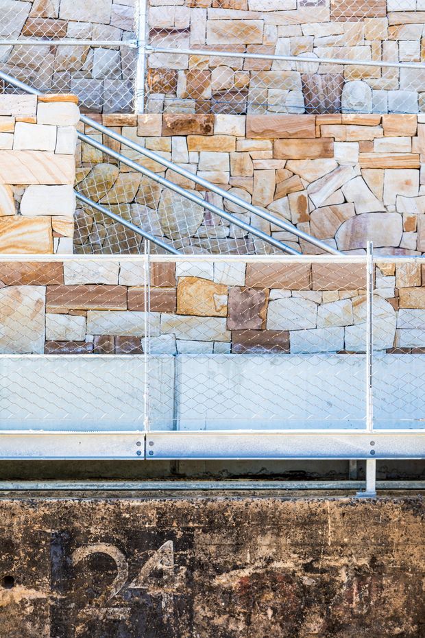 Close-up architectural detail of a stone-clad wall under construction, showing varied rectangular and irregular tan, cream, grey, and brown masonry behind temporary chain-link safety fencing and metal scaffolding rails. A weathered concrete base is visible at the bottom.