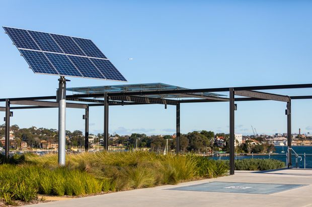 Exterior view of a contemporary waterfront structure with a large tilted solar panel array, steel posts and framing, and a flat open canopy. Native grasses and paved hardscape occupy the foreground, with water and a suburban shoreline visible beyond.