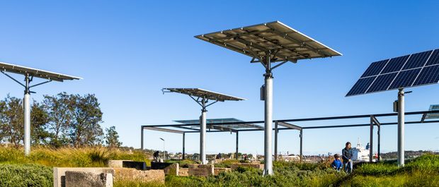 Exterior view of a solar energy installation with multiple elevated photovoltaic panel arrays mounted on steel support structures, set in a grassy landscape under a clear blue sky. Concrete blocks and a distant urban or industrial horizon are visible.