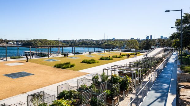 A bright waterfront public park and promenade with landscaped lawns, sandy-coloured paved open space, planted pergola structures, pedestrian paths, stairs, and a visible harbour shoreline with a distant urban backdrop.