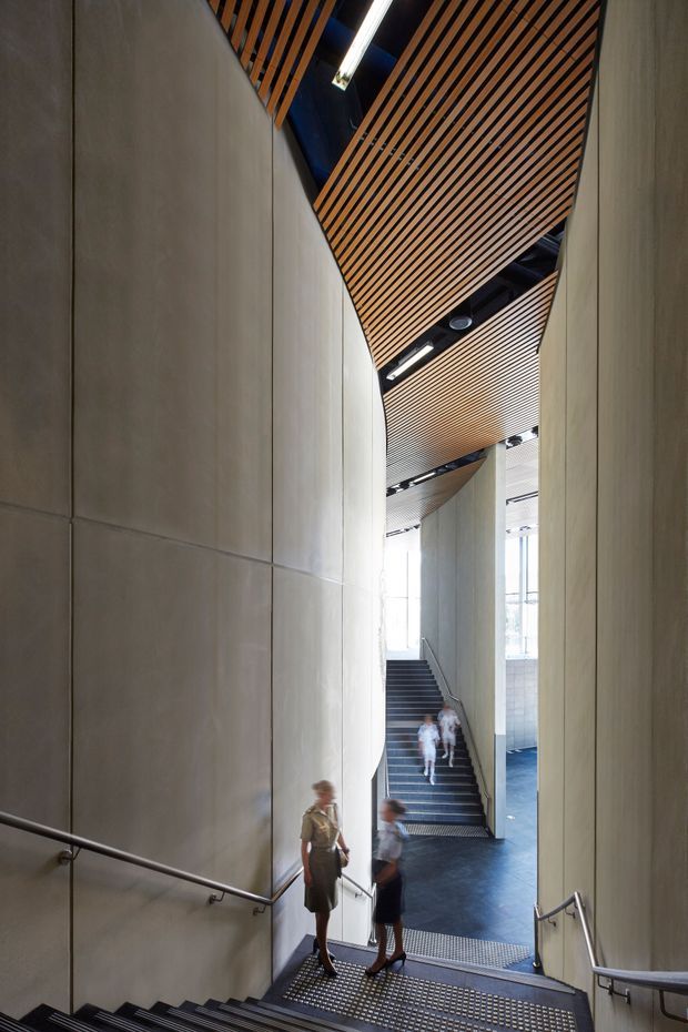 A tall, modern interior stairway or circulation space with broad concrete wall panels, dark metal stair treads and handrails, and a dramatic slatted timber ceiling. Multiple flights of stairs connect through a bright, multi-level public interior, with linear lighting and glimpses of glazing in the distance.