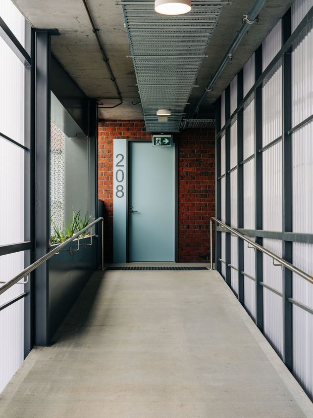 A narrow industrial-style access corridor or entry landing leading to a pale blue-grey door marked 208. The space has a smooth concrete floor and exposed concrete ceiling with cable trays, conduits, and simple round pendant lighting. Red brick forms the wall around the door, while dark metal framing, translucent ribbed wall panels, stainless-steel handrails, and a planted trough reinforce the utilitarian commercial aesthetic. An emergency exit sign is mounted above the door.