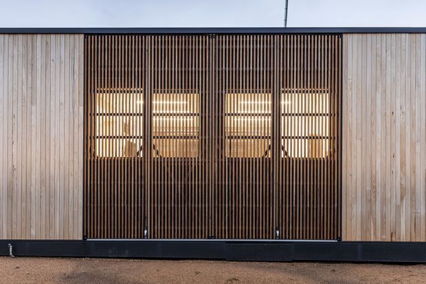 Front-on view of a contemporary single-storey facade featuring pale vertical timber cladding panels and a broad central screen of closely spaced dark timber battens. Warmly lit glazing and an entry or internal space are visible behind the slatted screen. A dark horizontal base and roofline frame the composition, creating a restrained, modern architectural exterior.
