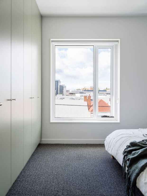 Bright, compact bedroom with a minimalist built-in wardrobe along the left wall, pale grey walls, white trim, dark grey carpet, and a partially visible bed with white bedding and a dark throw. A large white-framed window provides abundant natural light and views toward surrounding urban rooftops.