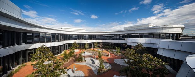 Wide exterior architectural view of a large contemporary multi-storey building arranged around a landscaped central courtyard. The building has curved wings, extensive dark glazing, white and light-grey metal-clad facades, projecting horizontal bands, and a flat roofline. The courtyard includes paved paths, planted garden beds, seating areas, and vivid terracotta-orange surfacing. Blue sky and clouds are visible above.